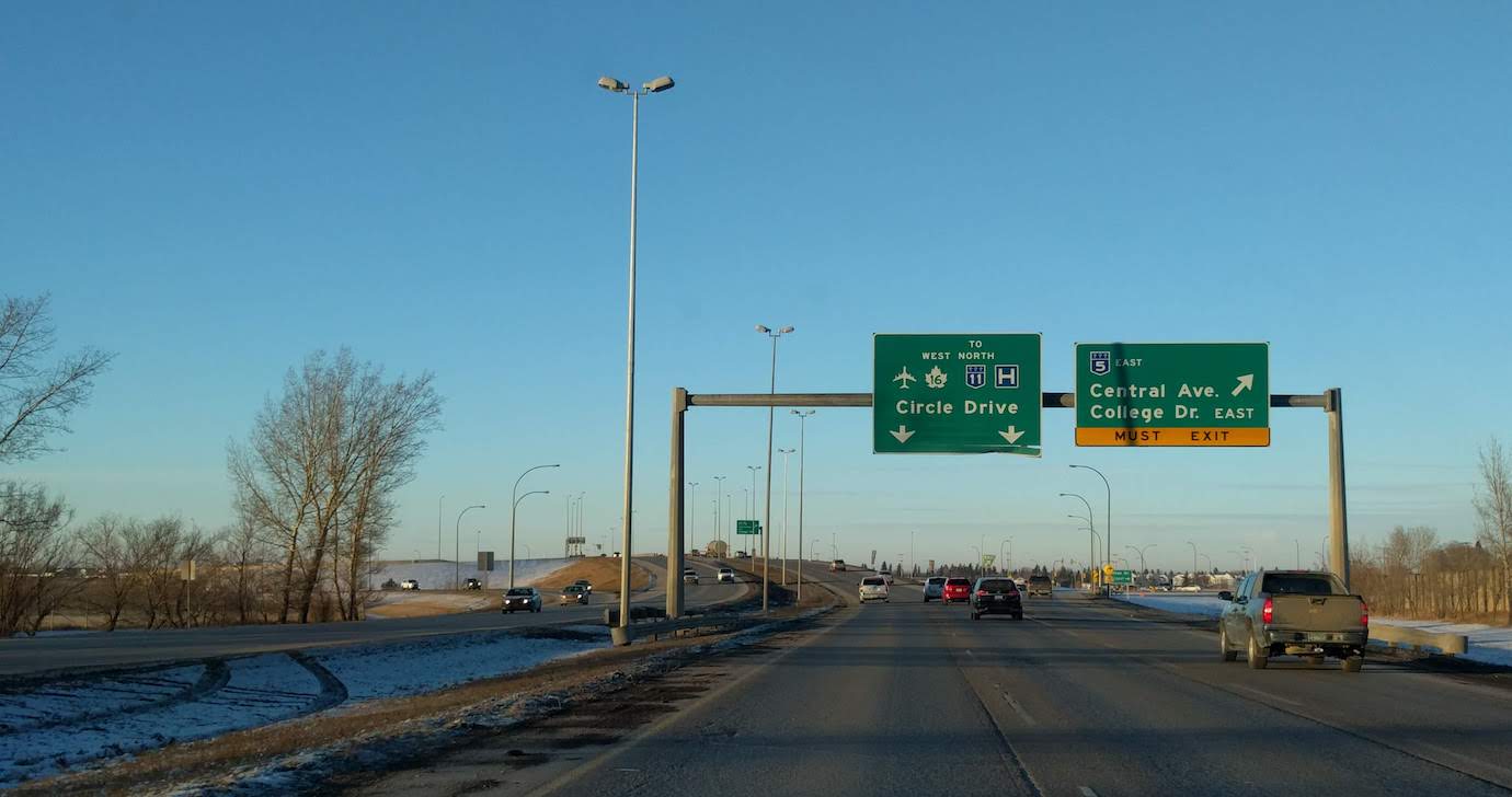 A photo of a highway overpass and offramp in Saskatoon, taken from the passenger seat of a car. The highway sign above says, “Circle Drive” ahead, “Central Avenue and College Drive East MUST EXIT” to the right