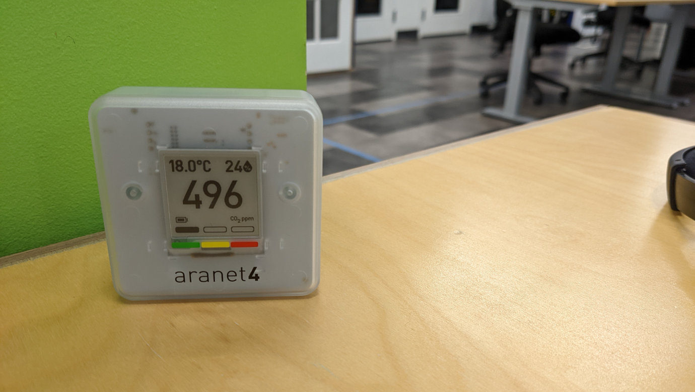 An Aranet4 CO2 sensor on a wooden desk: a small white square object with an e-ink screen. In the background there’s a green-painted wall on the left and other desks and chairs out of focus on the right. The e-ink screen says “496 CO2 parts per million”, a fairly low amount, with a green indicator below it (out of green, yellow, and red options) as well as the temperature (18 degrees Celsius) and humidity levels (24%) in the room.
