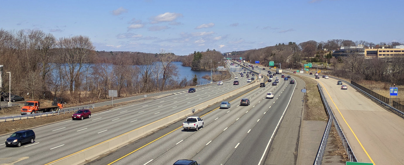 A multi-lane highway with medium traffic, on the outskirts of Boston, Massachusetts on a clear day. An industrial park and a lake are visible in the distance. (I clearly need more photos of Québec highway infrastructure!)