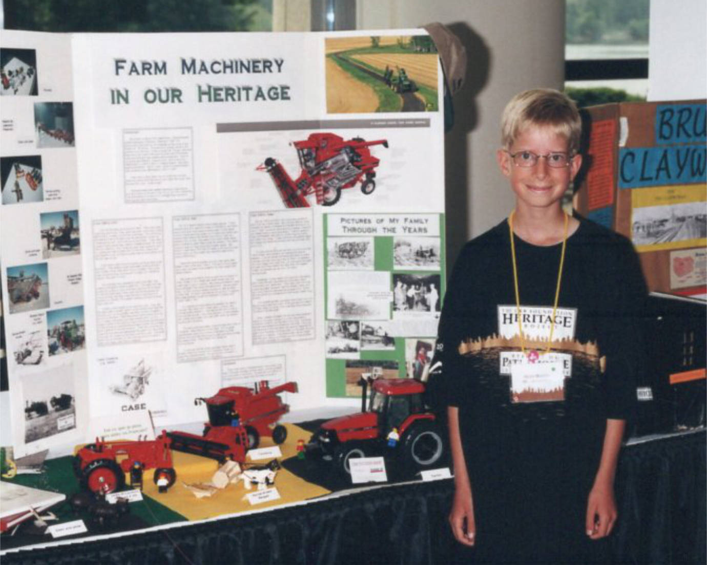 A small boy (the author) wearing an oversized “Heritage Fair” shirt, standing in front of a display titled “Farm Machinery In Our Heritage”, with several red model tractors and combine harvesters on a table in front of the display.