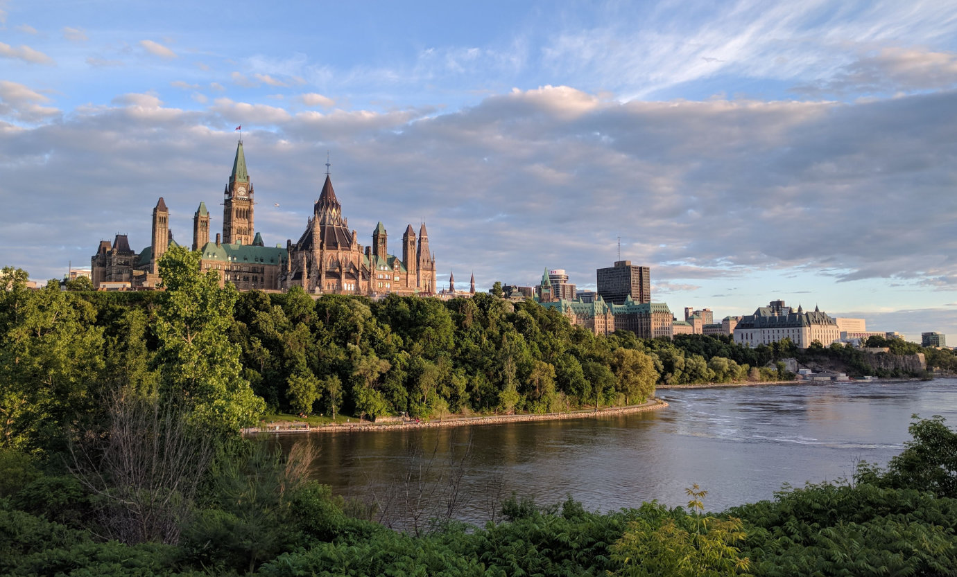 A photo of the Parliament buildings and Ottawa skyline, taken on a summer evening with trees and the river in the foreground.