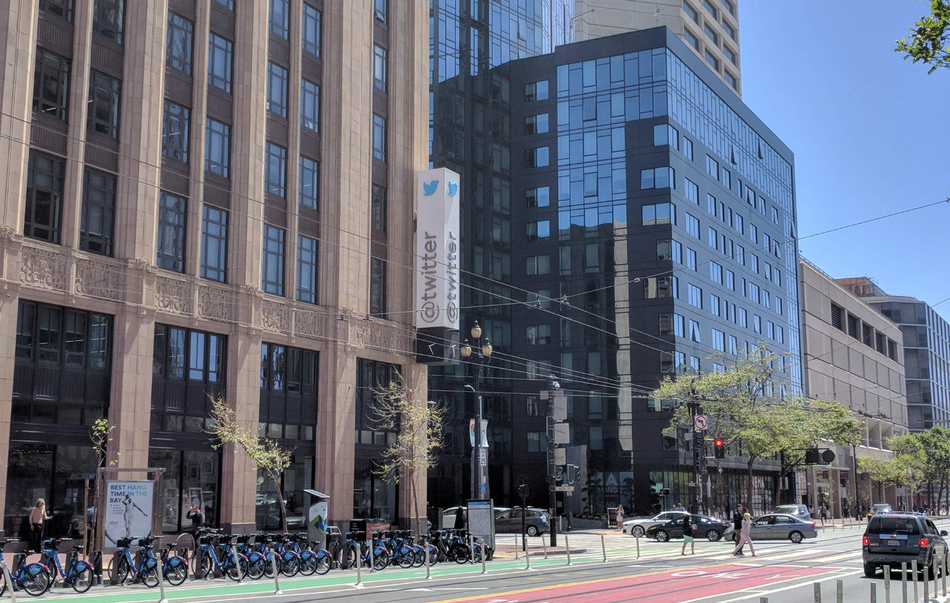 An angled photo of Twitter’s headquarters building, on a sunny day in San Francisco. A vertical sign saying “@twitter” is attached to the side of a brick-and-glass building. A long stand of municipal rental bikes, a bike lane, and tram tracks are visible in front, and to the right, another set of office buildings are visible behind an intersection with passing cars and pedestrians.