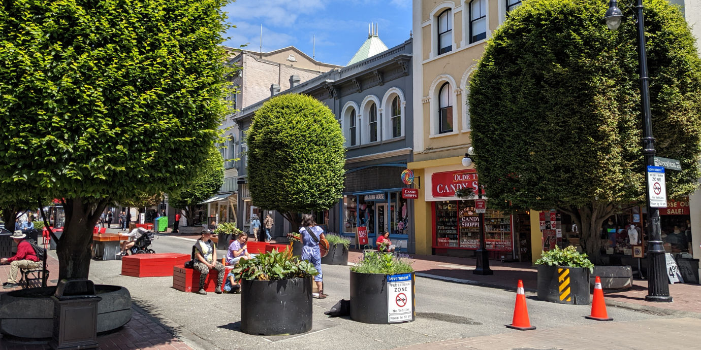 A sunny day on a pedestrian-only street in Victoria, with citizens and tourists sitting on red hexagonal wooden benches and a person playing a guitar. There are neatly-trimmed green trees on each side of the road, and a series of tourist-oriented shops in the background. A couple of signs say “Pedestrian Zone, No Vehicles, Noon to 10pm”.