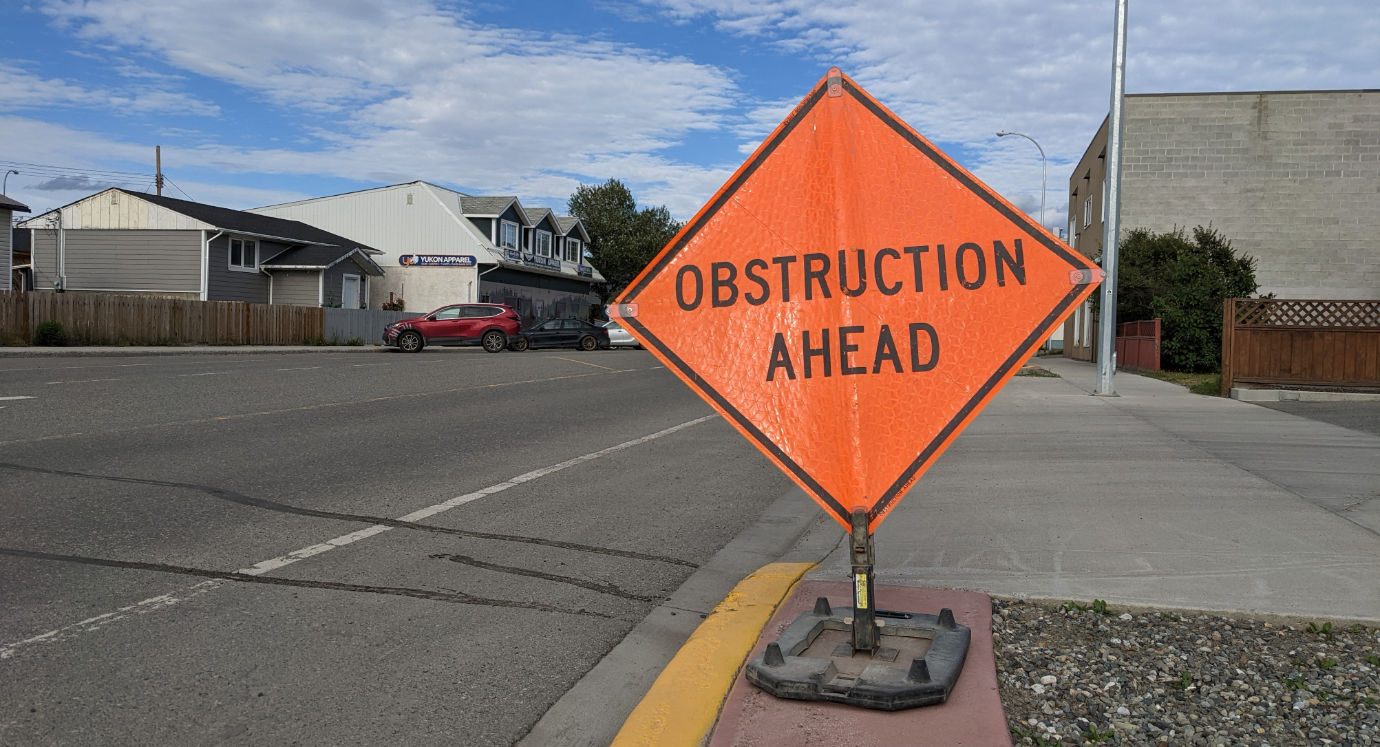 A bright orange construction sign on the edge of a downtown Whitehorse street, saying “Obstruction Ahead” in capital letters.
