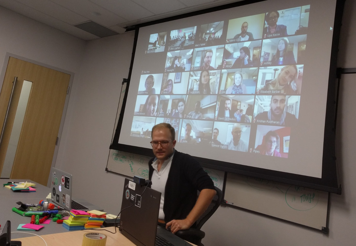 Alex Lougheed facilitating a multi-team all-staff meeting, sitting behind a table with two laptops and a large stack of Post-It notes and markers. In the background, an overhead screen shows a large Zoom call video grid, with 25 individual video feeds of various members of the Ontario Digital Service tuning in.