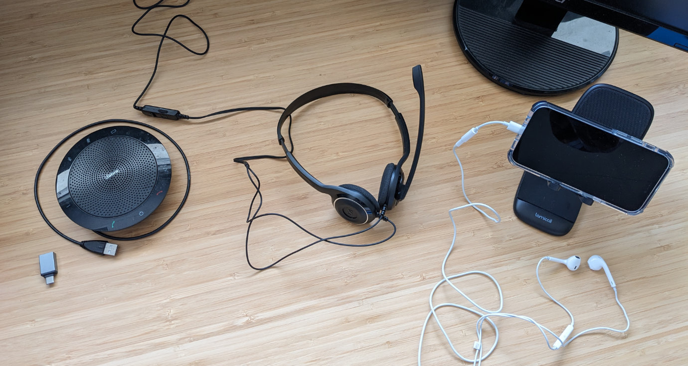 Three audio equipment options sitting next to each other on a wooden desk: a round Jabra 510 speaker/microphone puck; a Sennheiser PC-8 wired headset with thin arms; and an iPhone sitting on a portable phone stand with a Lightning-to-headphone jack adapter and wired headphones plugged into it. A computer monitor is partly visible in the top right corner of the photo.