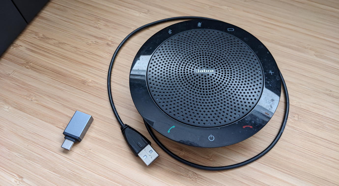 A round Jabra 510 speaker/microphone puck sitting on a wooden desk, with the USB cable partially unwrapped around it, and a small USB-A to USB-C adapter dongle sitting near it.