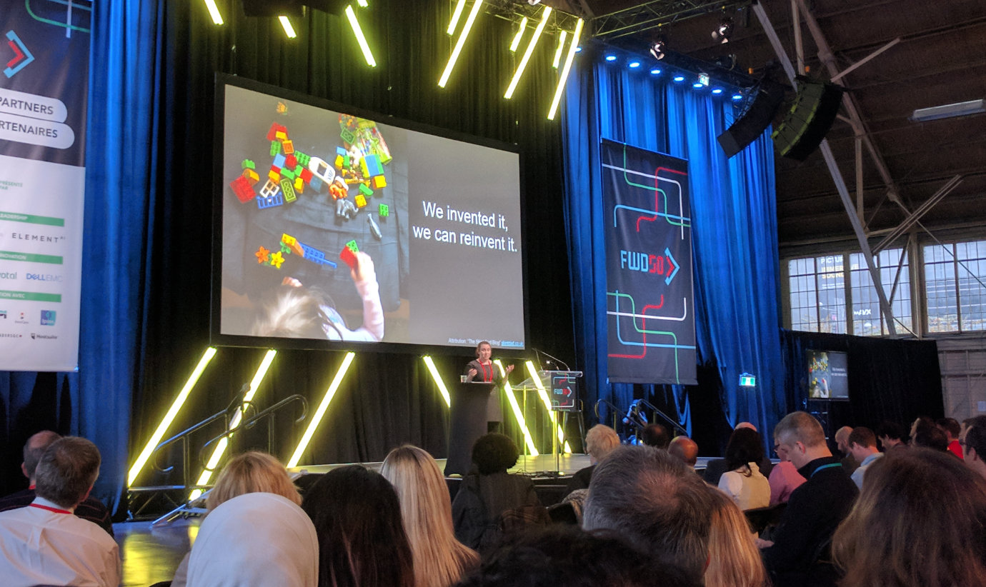 Pia Andrews on stage at the FWD50 conference, with banners and bright yellow angled lights behind her. A screen overhead shows her slides, which read “We invented it, we can reinvent it.” The backs of people’s heads are visible in the foreground. The windows and roof trusses of the Aberdeen Pavilion in Ottawa are visible in the distance.