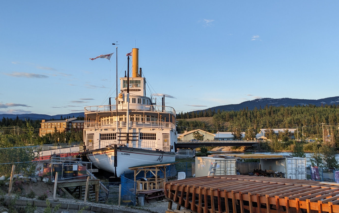 A front profile view of the S.S. Klondike paddlewheel steamer, on the shore of the Yukon River in downtown Whitehorse. In the background is the bridge across the river and an evening sky over the forest.