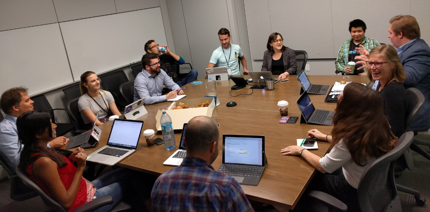 A room full of 12 people sitting around a boardroom table, smiling and laughing together. A bunch of Surface tablets and Macbooks are visible on the boardroom table, with some croissants in a box in the middle of the table.